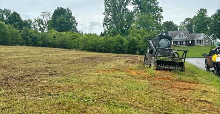 A wide view of land being prepared for development, featuring a skid steer loader on freshly turned earth, with residential properties in the background under a cloudy sky