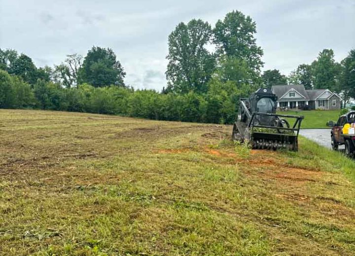A wide view of land being prepared for development, featuring a skid steer loader on freshly turned earth, with residential properties in the background under a cloudy sky